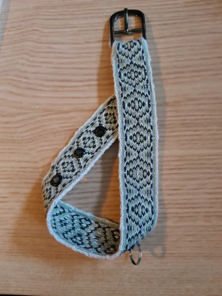 La sangle finie, avec une boucle et trois œillets noirs, un petit anneau de clefs pris dans la trame. Le motif est une répétition de polygones en noir, gris clair, sauge et blanc