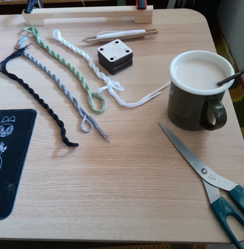 La mise en place, avec le dispositif de cadre arrimé à la table par deux serre joints, une navette remplie de coton blanc, quatre écheveaux de coton noir, gris, blanc et sauge, une tasse de thé et une paire de ciseaux