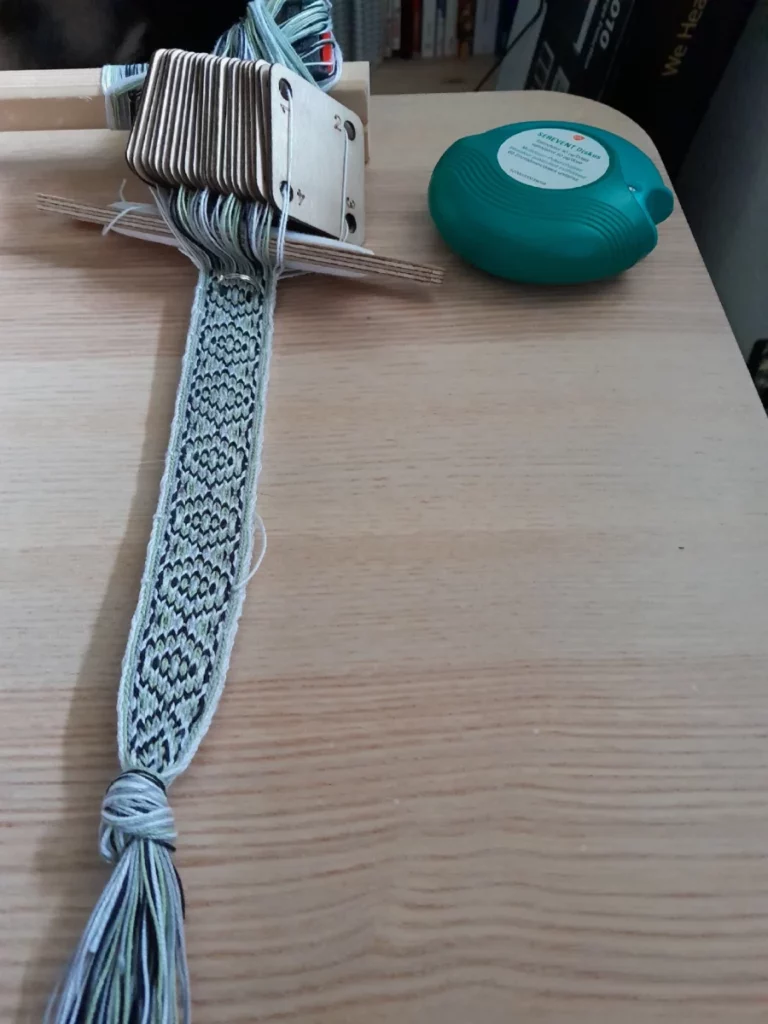 La sangle en cours de tissage est arrimée à une table par un petit dispositif en planchettes collées à la colle blanche, maintenues sur le bord par deux serre-joints. Les fils passent par une série de cartes en bois. La navette est retenue entre les cartes et la trame.