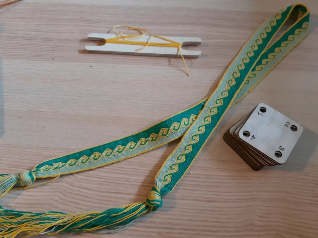 Une bande tissée aux tablettes, vert sauge d'un côté et vert forêt de l'autre, dessinant un motif évoquant les complications sur les amphores grecques, les deux moitiés séparées par un liseré jaune vif, et la bordure de la bande étant également de cette couleur. La bande est repliée en ruban sur la table, à côté d'une pile de fines tablettes en bois numérotées, et d'une navette en bois découpée à la CNC, encore un peu remplie de coton jaune.