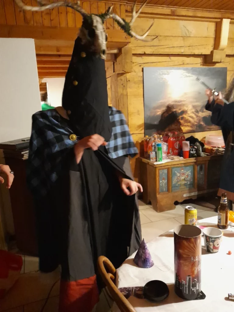 dans une grande salle au, murs de bois, une étrange créature à tète de renard ornée de bois de cerf danse près d'une table. elle a un très long cou affublé d'une pièce de tissu fermée par trois boutons dorés, dont dépassent quelques morceaux de sa fourrure. elle porte une grande cape grise et noire aux pans en flèches, terminée par une pièce colorée rouge et une blanche,  de chaque côté. Sa cape est surmontée d'un châle à carreaux bleus et noirs, tenu autour de son cou par une chaîne accrochée à deux camps dorés