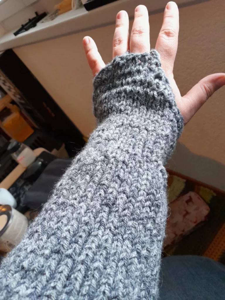 un manchon large en laines mélangées grises deux tons, part de la saignée pour s'arrêter au poignet,  couvrir le dos de la main uniquement.  il est fixé au majeur par une chaînette au crochet.