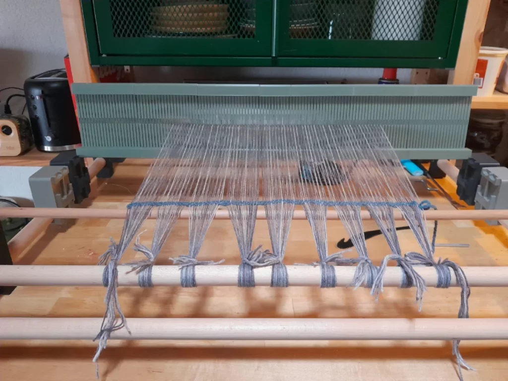 fixé à une table par deux serre-joints hors-champs, un métier à tisser est endilé sde laine grise chinée. le fil est juste tendu et noué, le travail attend d'être commencé