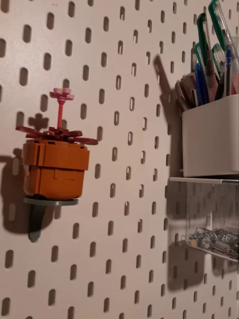 la plante accrochée à un pegboard, vue de côté et un peu en contre-plongée. Juste à côté il y a un pot plein de stylos et un petit casier transparent avec des punaises. C'est une drosifera stylisée, aux feuilles bordeaux composées de brosses à cheveux, avec une étamine d'un rose vif qui est en fait une rosace d'épaulettes.