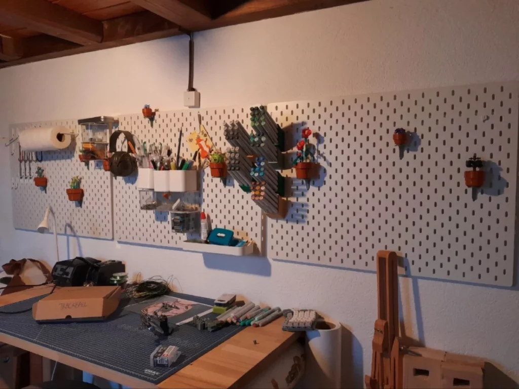 Une table à dessin avec une planche de découpe comme sous-main, devant un mur de pegboards sur lesquelles sont suspendues des stylos, un casque anti-bruit, des petites plantes lego sur leurs supports, des markers copic sur leurs supports, des petits bacs avec des punaises, des gommes et de l'encre, et d'autres machins...