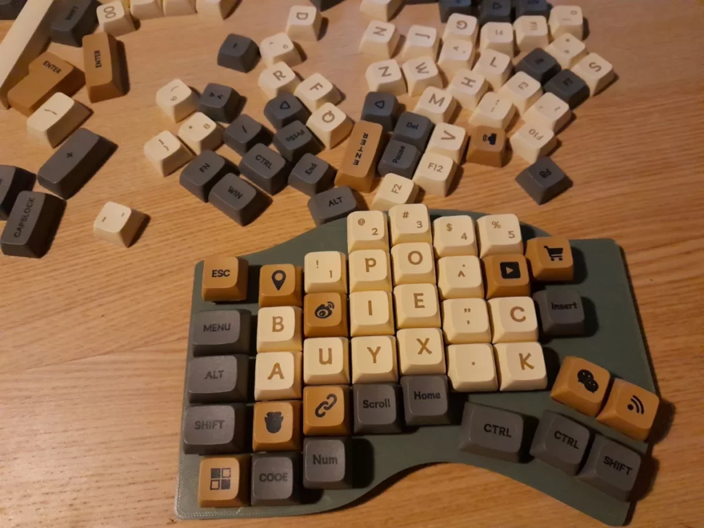 Un demi clavier split gauche. Le plate est couleur khaki bizarre - un pla recyclé et biodégradable - et les touches un mélange de couleurs grises,  crème et moka. La disposition est en bépo,  les touches sont souvent substituées par n'importe quoi. Il y a plein de touches du même set éparpillées derrière.