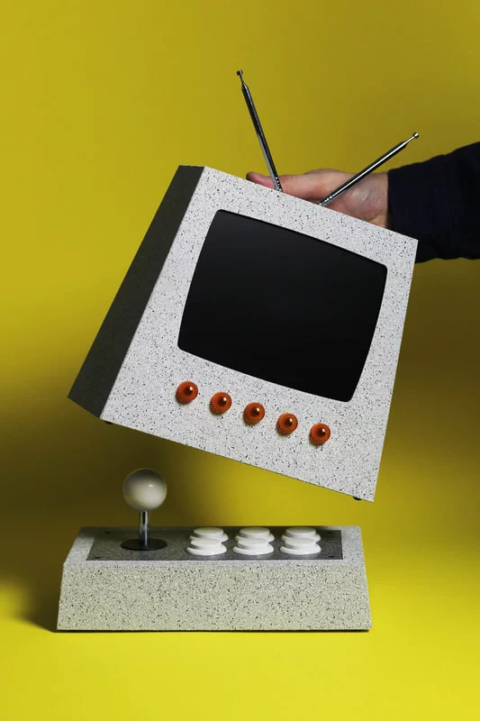 a tv set with a very retro-futuristic look, alike something one could see in a 60's sci-fi tv show. The TV set is pyramidal and glittery silver with five red buttons under the screen and two antennas. When you pull it up you discover it hides a little arcade stick with matching color and white caps