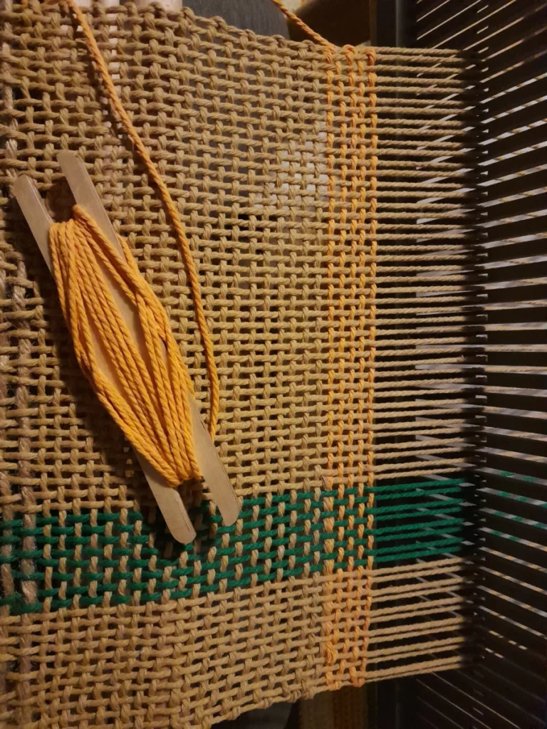 Un début de tissage très souple en gros coton pour macramé. La chaîne est majoritairement nature, avec une petite bande vert forêt légèrement à droite. La trame vient de changer du nature vers le jaune doré, amorçant un motif à carreaux.