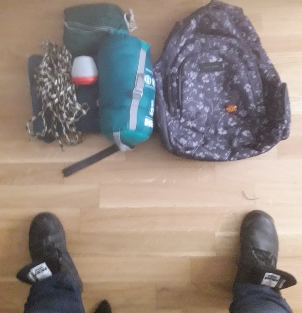 Je me tiens debout - rangers demi-ouvertes - devant mon sac à dos en préparation. Devant mes pieds on voit un sac à dos vide à côté d'un sac de couchage compressé, un hamac dans sa pochette, une petite lampe à recharge USB et un bon métrage de corde.