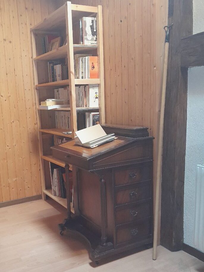 Le davenport vu de trois quart à côté d'une bibliothèque rudimentaire en pin massif, remplie de livres de poches. Les deux meubles sont dans l'angle d'une pièce aux murs recouverts de lambris