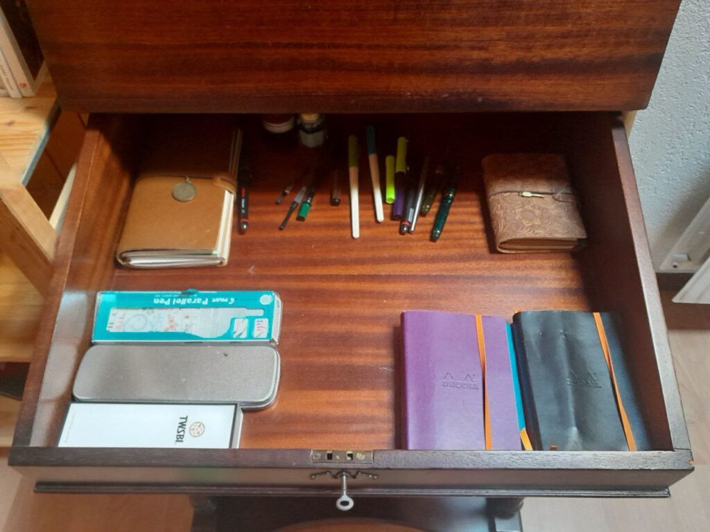 le coffre de l'écritoire est ouvert. Il laisse voir une petite pile de carnets lignés A6 en cours de remplissage, un traveler's notebook relié cuir miel taille standard dans le coin supérieur gauche, un traveler's notebook taille passeport relié cuir embossé floral dans le coin supérieur droit, au milieu il y a en vrac une petite collection de plumes et de réservoirs à piston, deux Jinhao, une Waterman, une TWSBI et deux parallel pen. Tout au fond il y a deux bouteilles d'encre, une sauge et une péridot. Dans le coin inférieur gauche, il y a la boite d'origine de la TWSBI, une boite d'origine de parallel pen, et une petite boite en fer blanc qui contient tous mes becs de plume et trois manches.