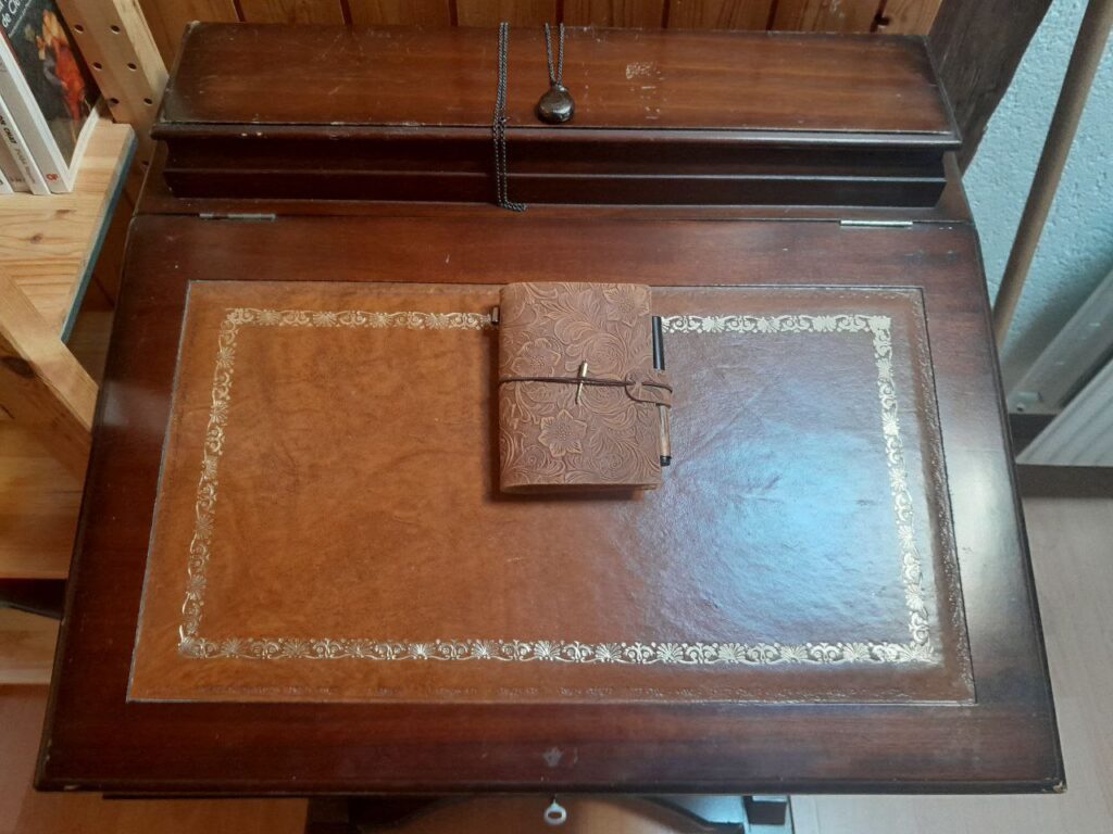 vue du dessus de l'écritoire. Tous les rangements sont fermés. Une petite montre à gousset noire sur chaine noire est posée sur le rangement supérieur. Le traveler's notebook taille passeport couverture cuir embossé floral trône au centre du sous-main cuir incrusté dans le plateau de l'écritoire. On peut voir les dorures sur les bords du sous-main en cuir dont on peut observer le profil texturé et très patiné. La lumière du vasistas crée un gros reflet de ciel bleu sur le bord droit du sous-main, révélant subtilement l'embossage discret sur le pourtour du sous-main.