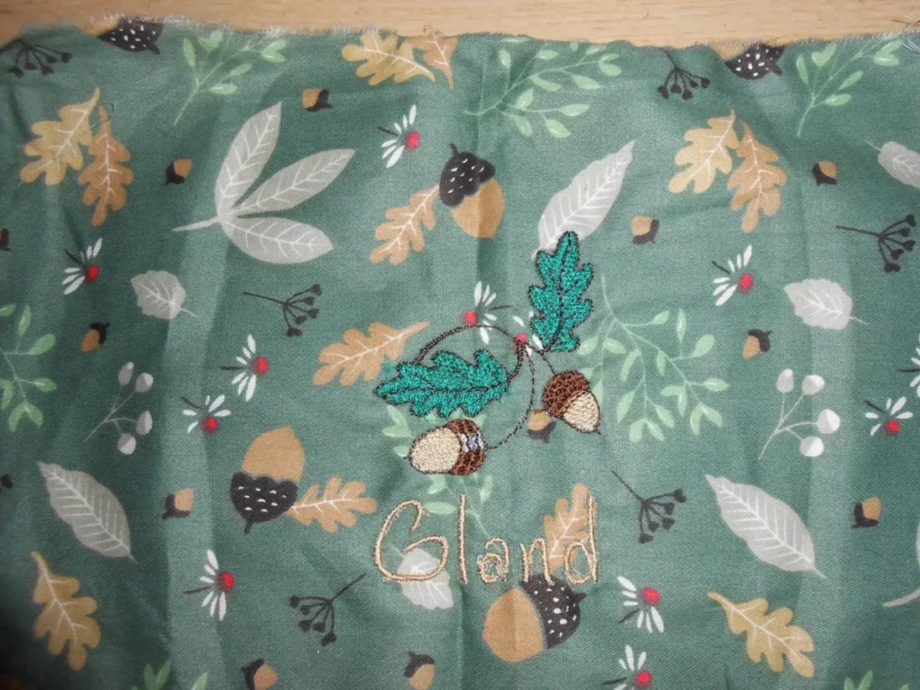 un carré de tissu vert avec un motif de feuilles mortes et de glands. Dessus j'ai brodé deux glands sur une branche avec deux feuilles, en-dessous c'est écrit "Gland" .