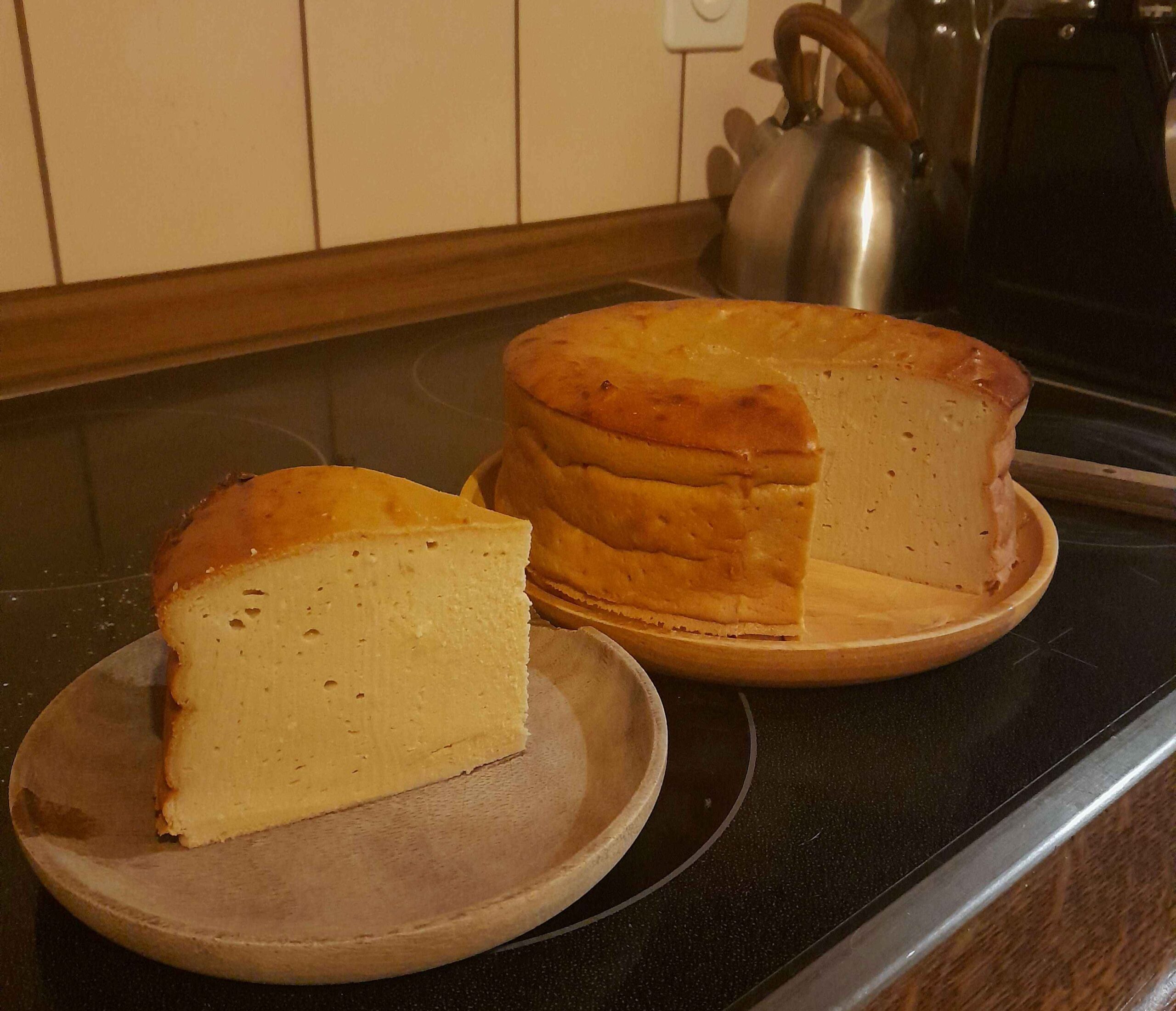 Un gâteau soufflé très clair, à la croûte dorée, trône sur une plaque chauffante. Il est cylindrique et bien haut. Il est déposé sur une assiette en bois clair. À côté, sur une assiette en bois foncé, il y a une part triangulaire du gâteau. La découpe est très nette, le gâteau se tient très bien. Il a l'air très moelleux.