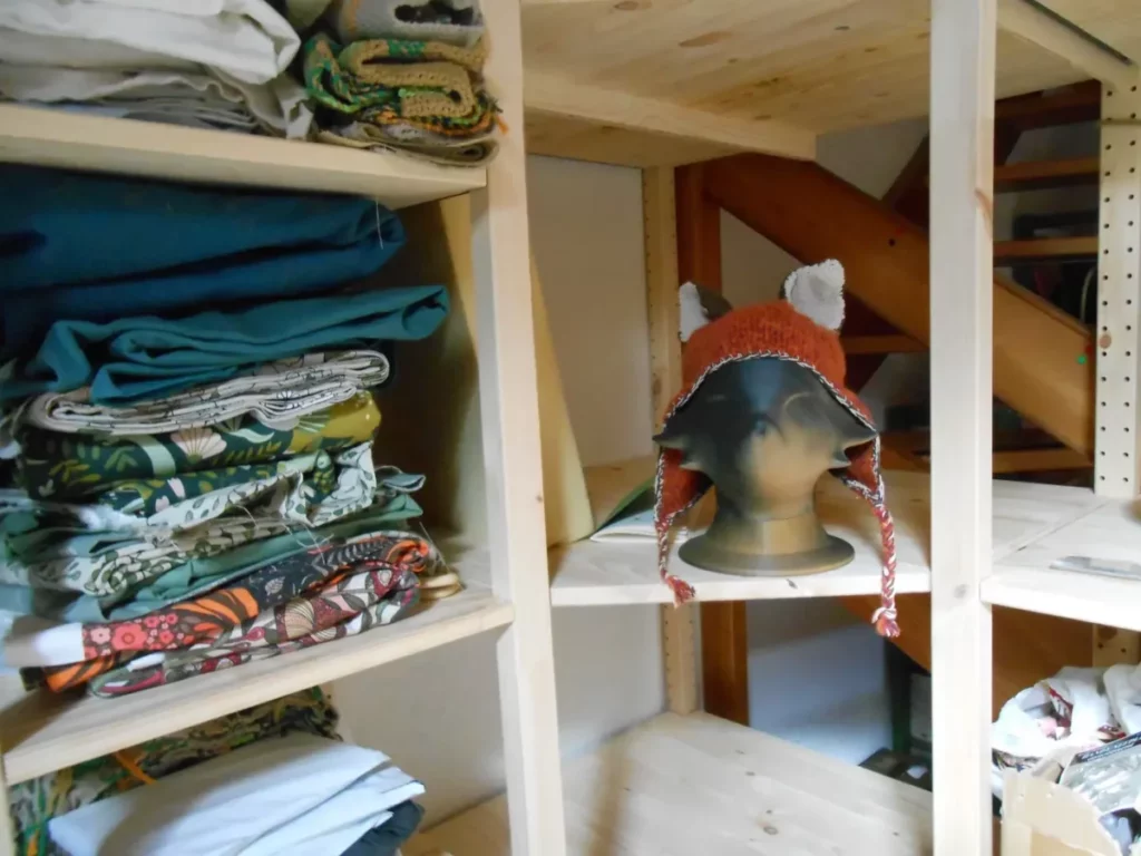 dans une étagère en bois, sur un rayon vide adjascent à un autre qui contient une grosse pile de tissus, se tient une tête de renard qui se décline d'une couleur un peu cuivrée sale jusqu'au noir. Elle porte un bonnet orangé à oreilles noires.