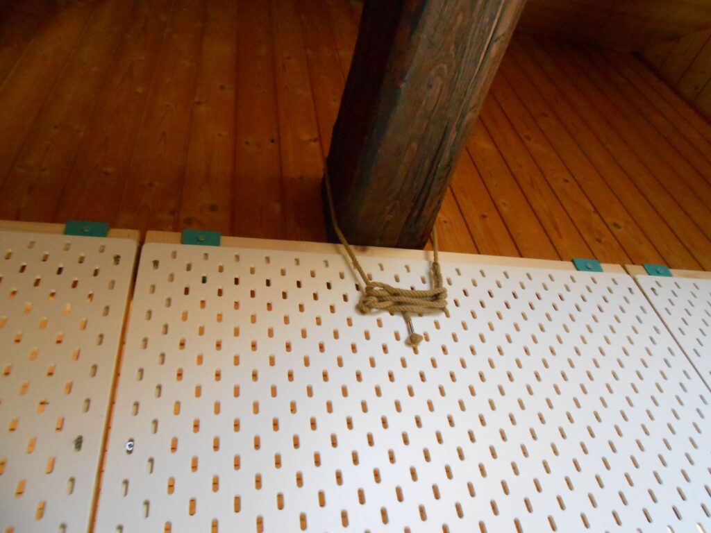 Gros plan sur une cloison, vissée au faux plafond de lambris, située sous une poutre environ à son milieu. On peut voir que les guides des cales de vissage sont aplatis. On voit aussi que la cloison est arrimée à la poutre à travers l'une des pegboards, à l'aide d'une corde.