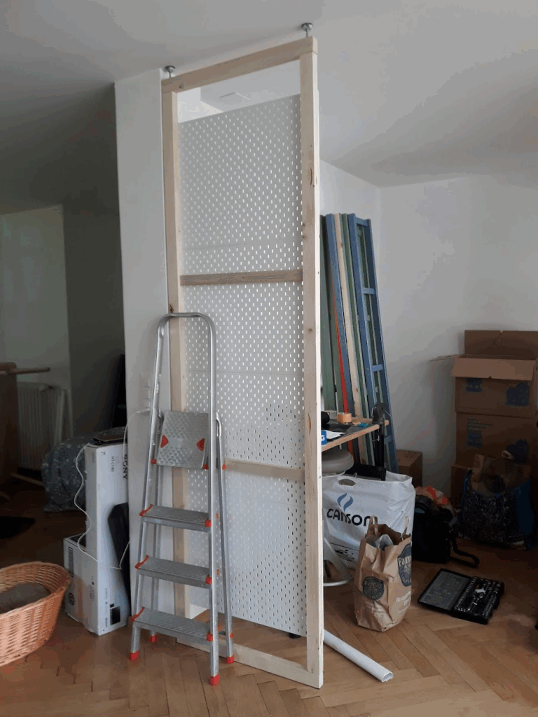 La première itération de cloison en pegboards. Un cadre en bois très simple, avec deux barres transversales pour la stabilité. Il est fixé dans l'espace entre le sol et le plafond par un système de vérins composés de vis, de boulons autobloquants et de disques à pas-de-vis, complétés par des petits tampons de caoutchouc pour amortir les chocs.