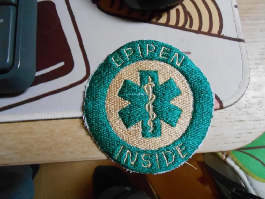 un patch avec une croix médicale avec un serpent au centre et un cercle externe sur lequel il est écrit "EPIPEN INSIDE". Il est à mes couleurs, beiege pour le fond du cercle interne, du serpent et du texte, vert pour la choix et le cercle externe.