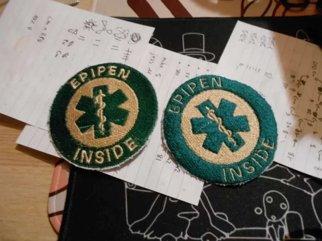 Deux patches ronds, verts sur fond bbeige, avec une croix médicale ornée d'un caducée, entourée du texte "EPIPEN INSIDE". Celui de droite est le proto avec ses défauts : texte trop fin, cercle externe trop épais. Celui de gauche est la correction : texte plus gras, ainsi que le caducée qui pop bien maintenant, et le cercle externe bien plus fin.