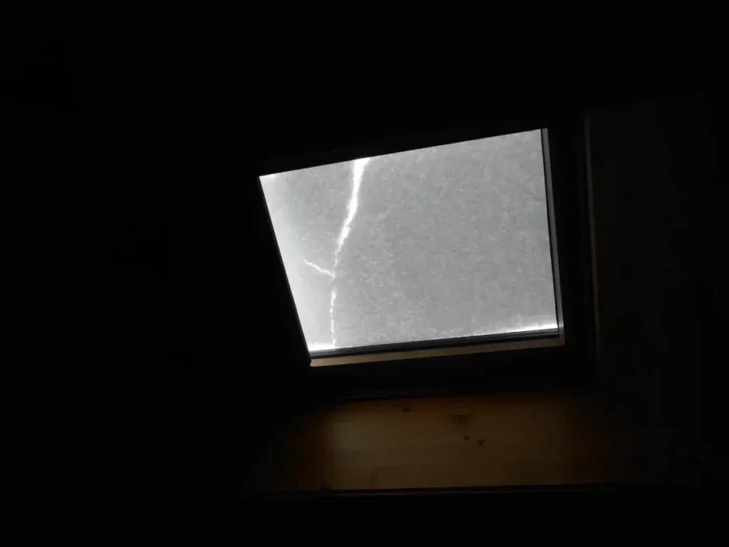 un vélux recouvert de neige. Le bloc de neige est fissuré en haut à gauche. C'est un petit carré très lumineux dans une pièce très sombre.