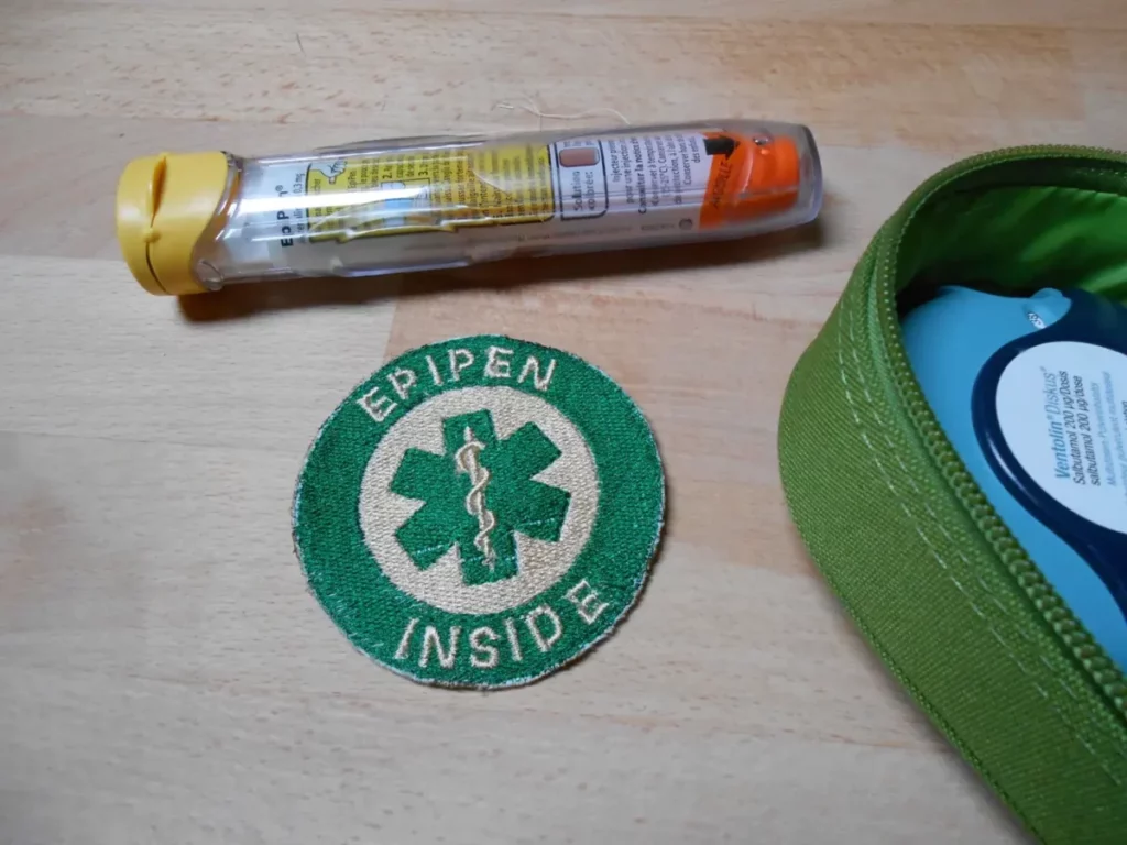 Sur une table, un patch est déposé entre une trousse de secours où on peut voir un bout d'un discus de ventolin et un epipen. Le patch est circulaire. Au centre il y a une croix médicale à six branches ornée d'un caducée, verte sur fond beige. Un anneau vert la borde, sur lequel il est inscrit en lettres capitales "EPIPEN INSIDE".