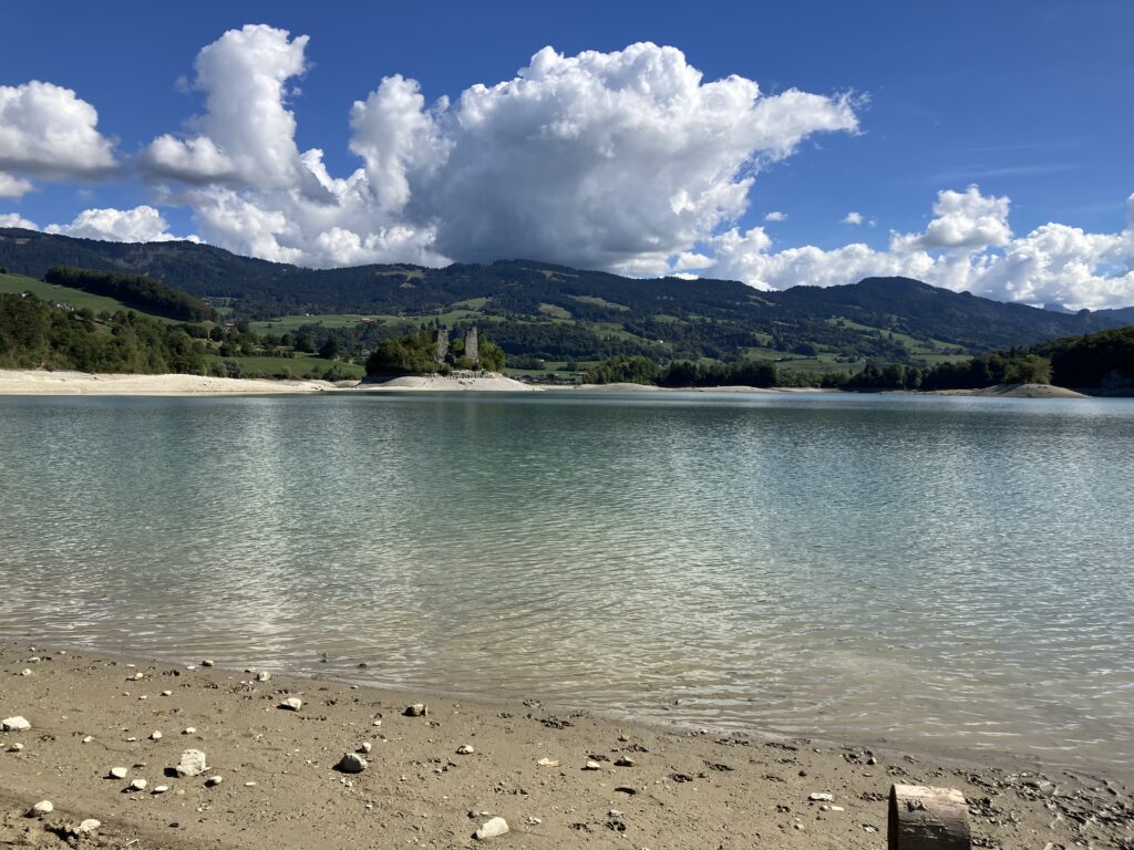 Une photo d'un lac d'eau limpide au mlieu des montagnes - on ne voit pas le Moléson, les pics environnants sont plutôt bas. Au centre de l'objectif on voit une presqu'île reliée à la plage par une bande de remblais en pierres. Au centre de l'île se dressent deux tours médiévales. Quelques nuages agrémentent un grand ciel bleu.